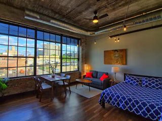 A chic industrial-style loft apartment with exposed ductwork, a large window providing city views, and a blue patterned bedspread
